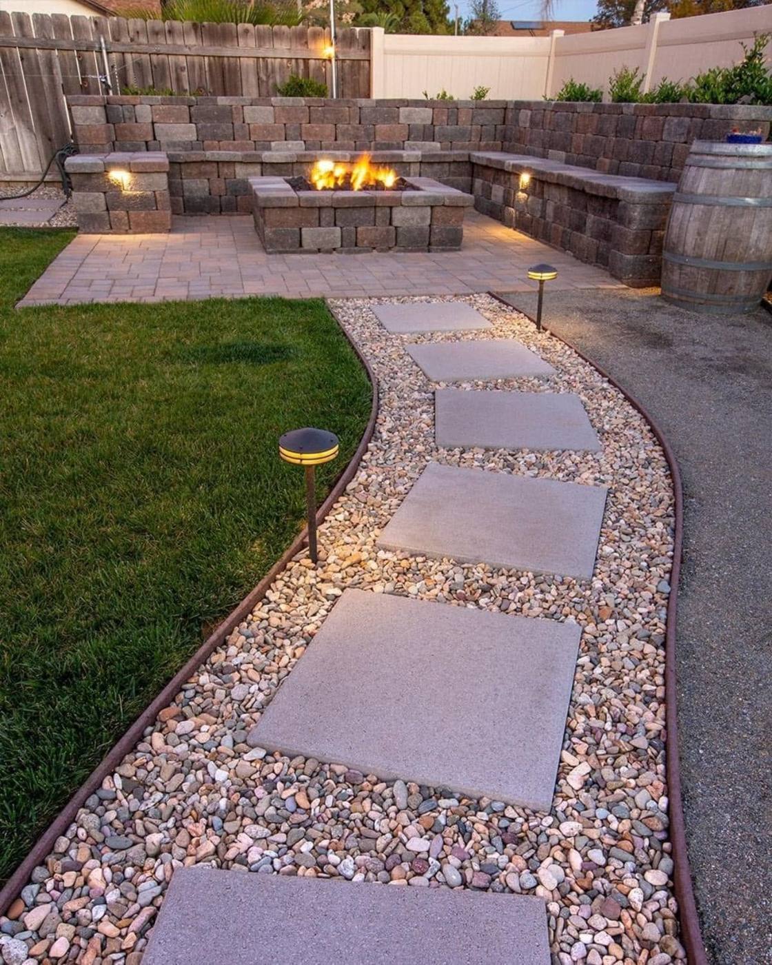 Backyard at dusk: a curved path of large stepping stones set in river gravel and lit by path lights, leading past new lawn to a block fire pit and seating wall
