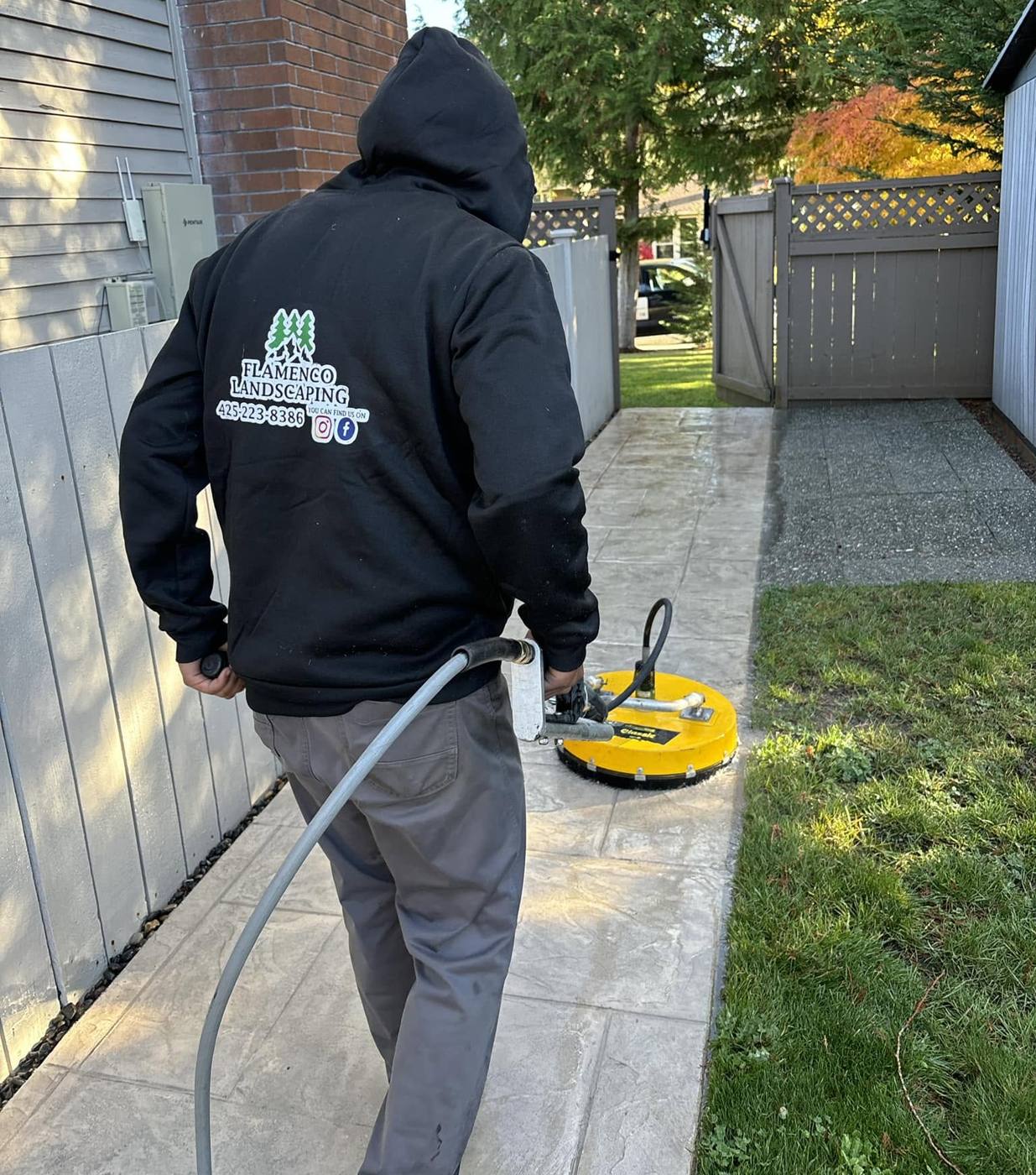 Crew member in a Flamenco Landscaping hoodie pressure-washing a paver walkway with a surface cleaner